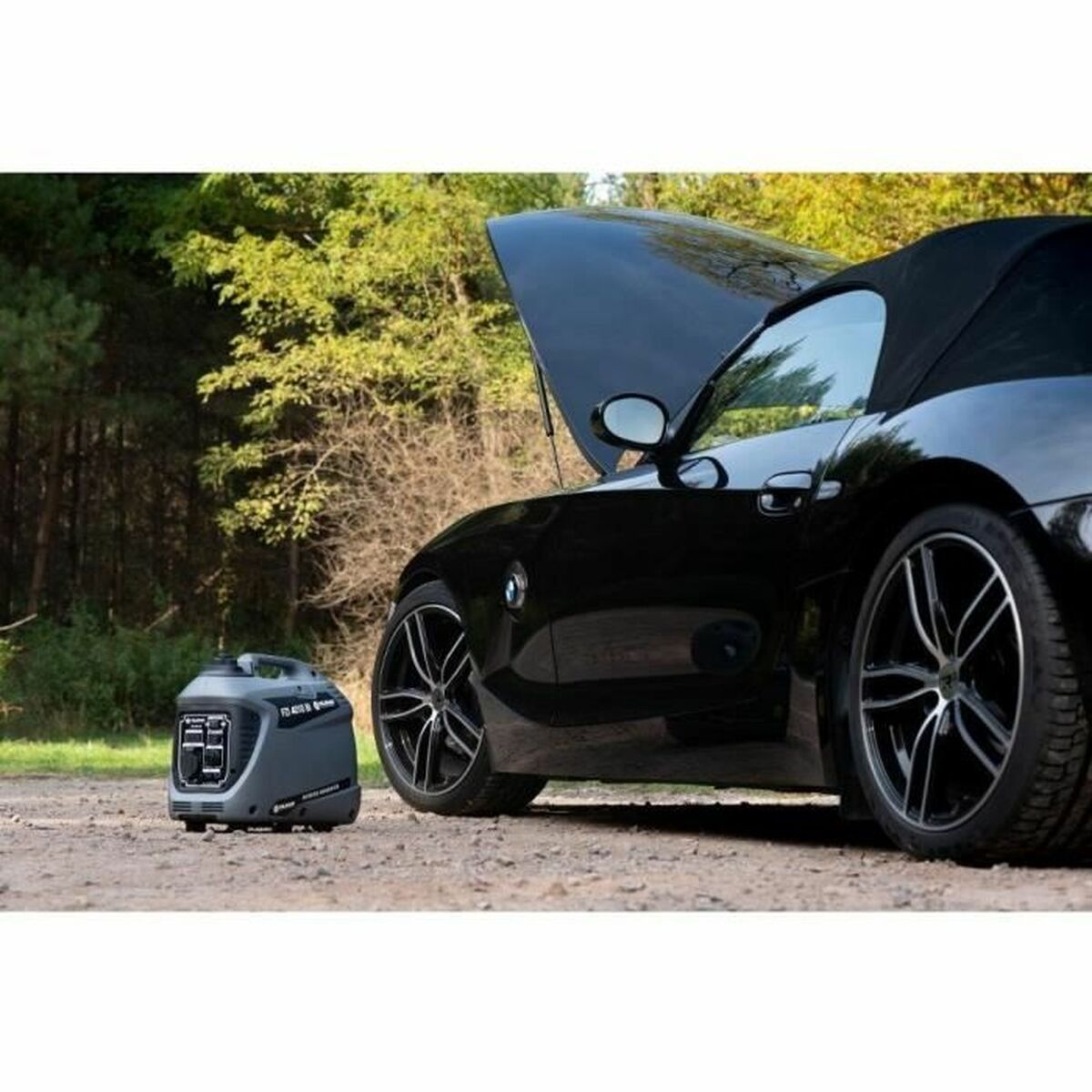 Fieldmann FZI 4010-Bi inverter aggregaat naast een zwarte cabrio met open motorkap op een grindpad in een bosrijke omgeving